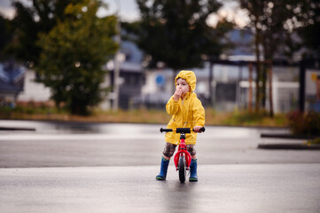 Young boy in a yellow raincoat and blue boots, riding a red balance bike on a wet street, enjoying a rainy day with a curious expression.