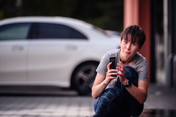 Woman crouching on a wet pavement, holding a smartphone, wearing casual workout clothes, with a white car in the background.