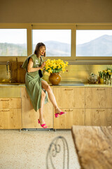 Young and elegant woman sitting on a kitchen table top with a flowers in vase, decorating her cozy and style interior of a house in mountains