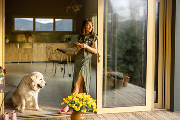 Young woman stands at an open window on a terrace with her cute white dog nearby, spending leisure time at home. Love, rest and friendship with pets