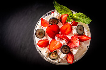 Strawberries with whipped cream and cottage cheese cream, mint and blueberries. Dessert in a glass on a black mica board. Copy a space