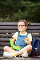 Cute little schoolgirl eating from lunch box outdoor sitting on a bench. Food for kids.