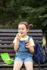 Cute little schoolgirl eating from lunch box outdoor sitting on a bench. Food for kids.