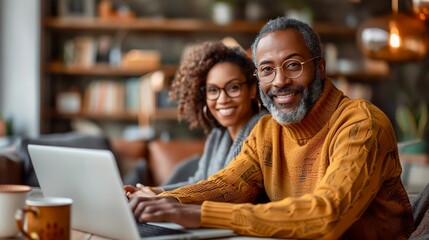 A happy couple is sitting at their kitchen table and looking at their laptop. The woman is smiling and the man is looking at the camera.