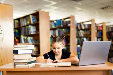 a young girl in black headphones sits at a table on which there is an open book, in a library room, studying.