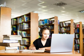 a young girl in black headphones sits at a table on which there is an open book, in a library room, studying.