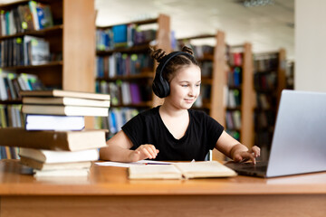School library. Schoolgirl studying online using laptop, taking notes on paper. Teenage schoolgirl wearing headphones undergoes online learning. Distance learning concept.