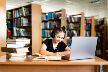 School library. Schoolgirl studying online using laptop, taking notes on paper. Teenage schoolgirl wearing headphones undergoes online learning. Distance learning concept.