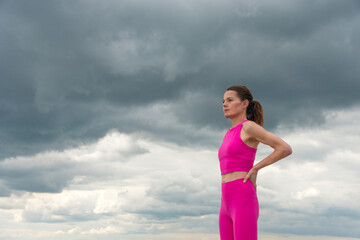 Sporty woman standing resting with hands on hips wearing pink sports clothing against a dramatic sky.