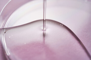 Serum or cosmetic oil flows into a transparent bowl on a purple background.