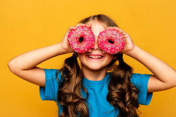Portrait of a little smiling brunette girl with two appetizing pink donuts in her hands, closes her...