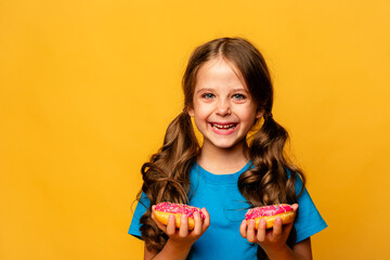 Portrait of a little smiling brunette girl with two appetizing pink donuts in her hands on a yellow...