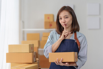 Woman in apron holding a cardboard box with a finger to her lips in a quiet gesture in a room...