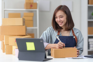 Young woman wearing an apron, packing a box while working from home with laptop and stack of...