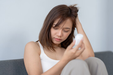 Young woman with a headache looking at a pill bottle, sitting on a gray sofa, feeling unwell, and holding her head.