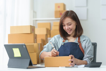 Smiling woman preparing packages for shipping in a home office, surrounded by boxes and working...