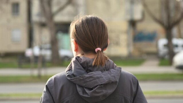 Rear view of adult brunette female standing on the street and looking at traffic and cars passing by