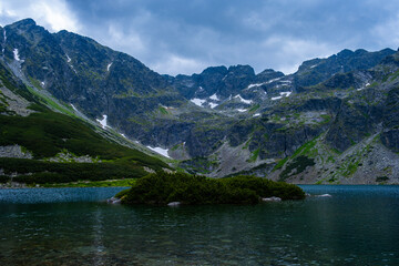 A landscape of rocky mountains and a lake with a small single island with green bushes in cloudy weather