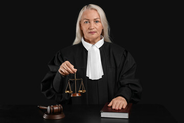Mature female judge with scales of justice and book at table on black background