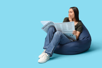 Fototapeta premium Shocked young woman in beanbag chair with newspaper on blue background