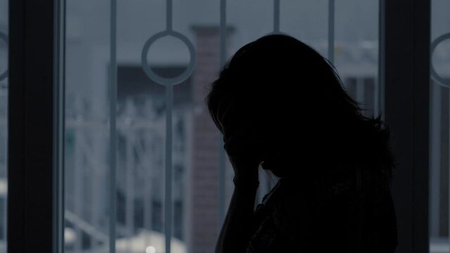 Backlit silhouette of a woman in her 40s with long dark brown hair, bringing her hands to her face in despair. The window behind her shows a gloomy day, reflecting her somber mood.