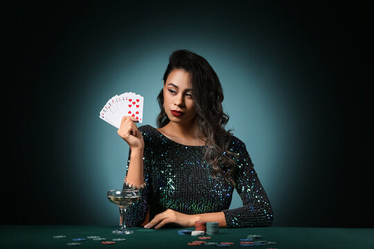 Beautiful young African-American woman sitting at poker table with playing cards, chips and glass of champagne on dark green background