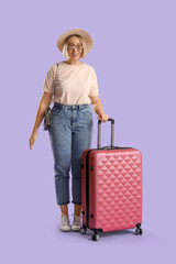 Young female tourist with suitcase on lilac background