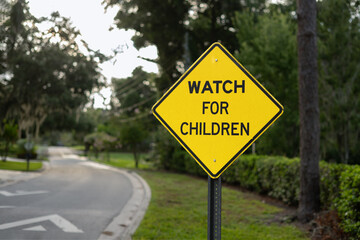 WATCH FOR CHILDREN, a yellow and black diamond sign surrounded by trees and a dirt road in the background.