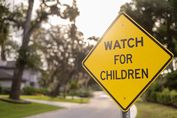 Close-up WATCH FOR CHILDREN yellow sign at sunrise surrounded by foliage with a dirt road in the background, horizontal