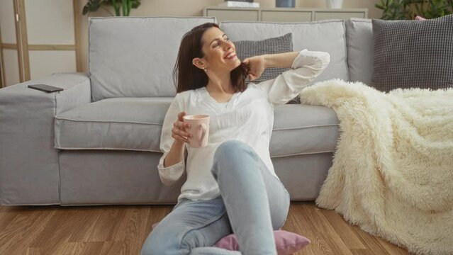A young hispanic woman in a white blouse, enjoying a relaxed moment with a coffee mug while sitting on the floor of her cozy living room in an urban setting.