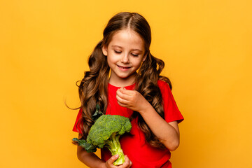 Happy smiling little girl in red t-shirt standing with green raw broccoli over yellow background. Healthy food for children concept