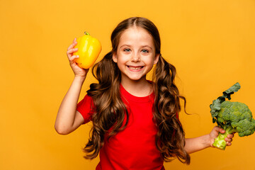 Happy little girl standing with green raw broccoli and yellow pepper over yellow background. Healthy food for children. Little girl with eco vegetables, isolated, space for text, concept organic food