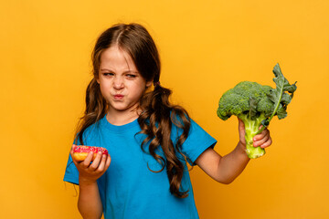 Little cute kid girl refusing to eat healthy vegetables broccoli on yellow background. Children do...