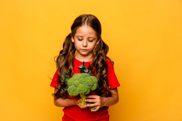 Little cute kid girl refusing to eat healthy vegetables broccoli on yellow background. Children do...