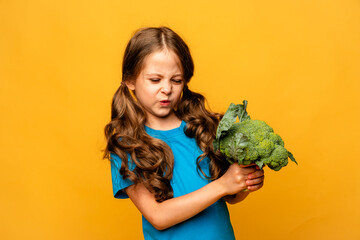 Little cute kid girl refusing to eat healthy vegetables broccoli on yellow background. Children do...