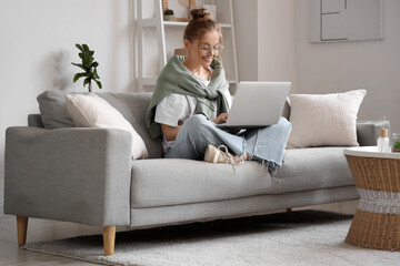 Fototapeta premium Young woman in eyeglasses sitting on sofa and chatting with laptop in living room