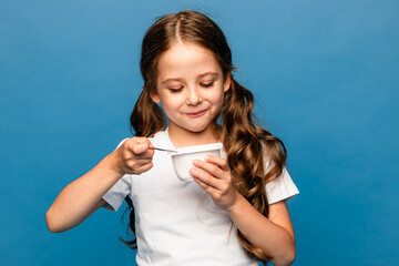 Cute little brunette girl in white t-shirt eating tasty yogurt on light blue background. Healthy...