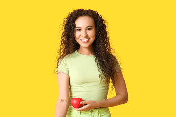 Young female African-American blood donor with applied medical patch and heart on yellow background