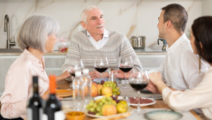 Calm smiling aged man enjoying family dinner with wife, adult daughter and friendly son-in-law, sharing stories and laughing over wine and appetizers in cozy kitchen