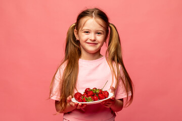 Portrait of a little blonde girl in pink t-shirt eating strawberry against pink background. GMO free food. Kids eat fruit outdoors. Healthy snack for children. summer concept. High quality photo