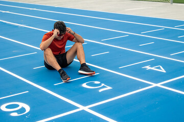 Exhausted Male Runner with Sunglasses Sitting on the Ground at Finish Line of Blue Running Track