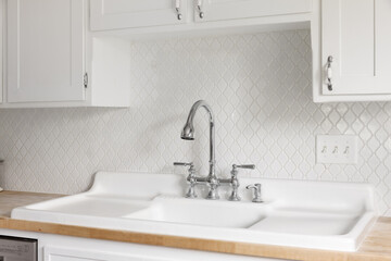 A kitchen faucet detail with a large white farmhouse sink with double drainboards, arabesque backsplash tiles, and natural butcher block countertop.