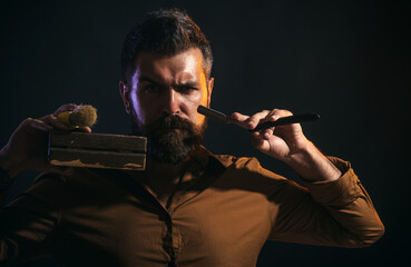 Portrait of brutal bearded man in stylish shirt with straight razor, wooden box and shaving brush. Barbershop service. Professional barber ready to shave you. Hairdresser with barbershop accessories.