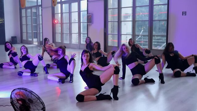A large group of female dancers are learning to dance in high heels in an evening class. Eleven girls are dancing on the floor in a doggy style pose.
