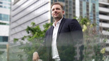 Caucasian business man hold mobile phone and looking at skyscraper view. Skilled male leader standing rooftop in modern building. Caucasian executive manager look at phone while rest his eyes. Urbane.