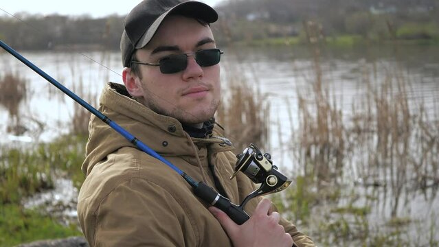A young man, in a blue jacket, glasses, with spinning rods in his hands, sits in nature near the river, enjoying the silence and dreaming of a good catch. Slow motion.