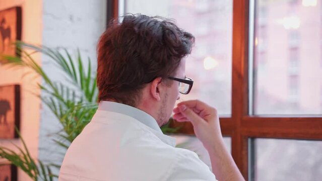 Man doctor with glasses in white coat near window, thoughtfully rubbing chin, pompously straightening hair with hand, turning head arrogantly Narcissistic professor, egoist gesture of superiority