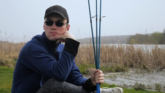 A young man, in a blue jacket, glasses, with spinning rods in his hands, sits in nature near the river, enjoying the silence and dreaming of a good catch. Slow motion.