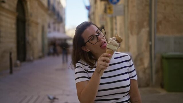 Young hispanic woman enjoying a large ice cream cone on a sunny day in the charming town of lecce, puglia, italy, wearing a striped shirt and glasses while expertly savoring the treat.