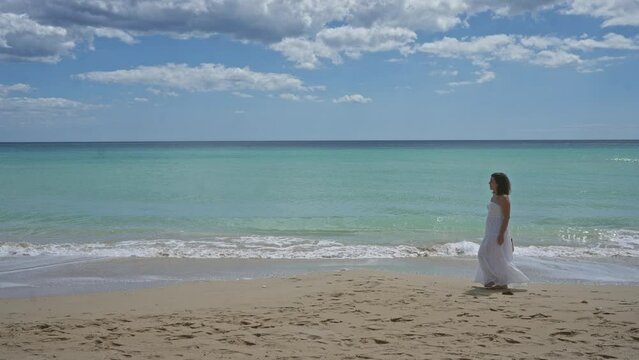 Beautiful young hispanic woman walking on the sandy beach of pescoluse in salento, puglia, italy, with clear blue waters and a cloudy sky in the background.